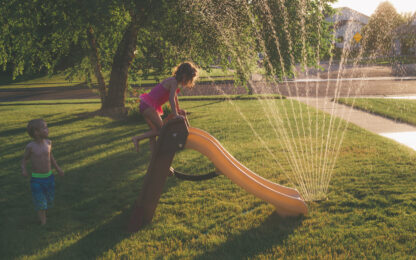 Little boy and little girl play outside and run through a lawn sprinkler and slide down a slide on a hot summer evening.